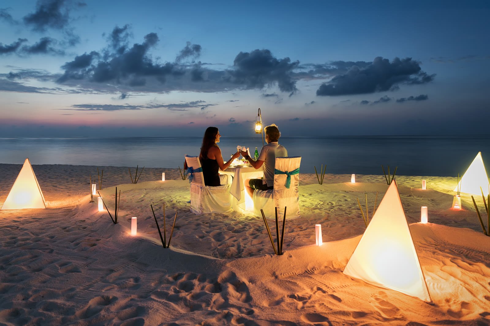 Couple toasting at a lantern-lit table on the sand at twilight