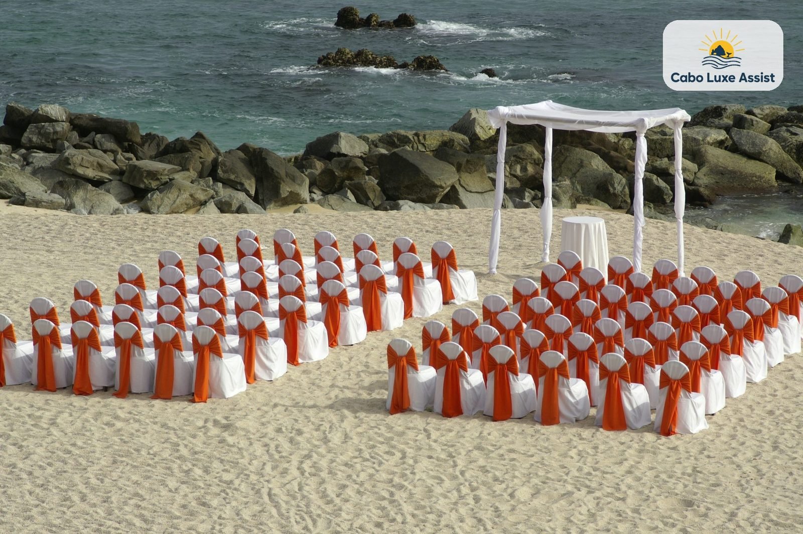 White chairs with orange sashes on Cabo beach sand facing a white canopy at the rocks, the Pacific behind